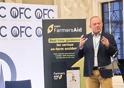 A panel of five people sits on stage during a conference discussion. They are seated in a row on chairs, dressed in business attire, and holding notes or microphones. Behind them, large illuminated co