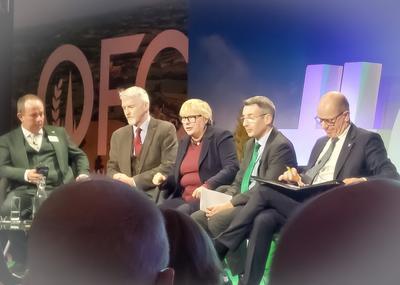 A panel of five people sits on stage during a conference discussion. They are seated in a row on chairs, dressed in business attire, and holding notes or microphones. Behind them, large illuminated co